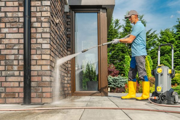 A man wearing yellow boots and gloves is using a pressure washer to clean the brick wall of a house on a sunny day. Lush greenery surrounds the area.